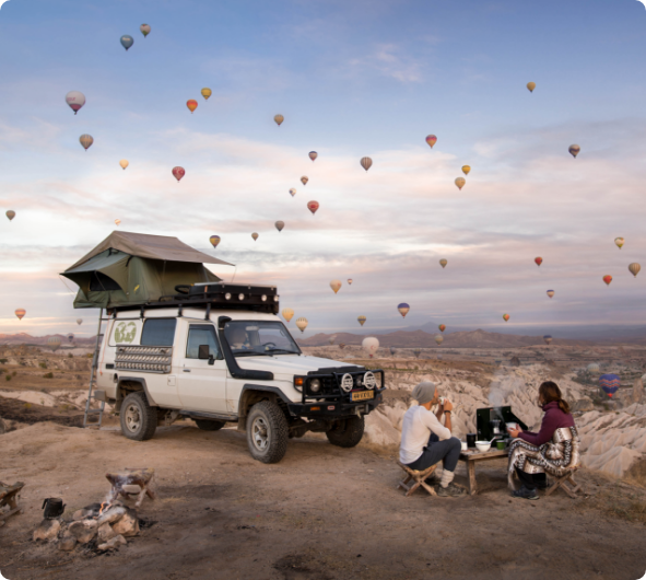 Jeep mit Dachzelt und zwei Personen sitzen an kleinem Tisch. Im Hintergrund eine tolle Landschaft mit hunderten von Ballons in der Luft